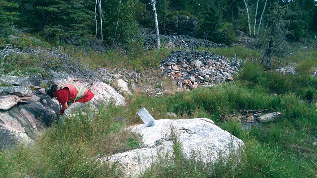 Prospector Robert Freeman sampling in 2016 at New Age Metals’ Lithium One lithium property in Manitoba. The sample bag sits on an outcrop of the Spodumene-Lepidiolite Zone. Photo by Carey Galeschuk.
